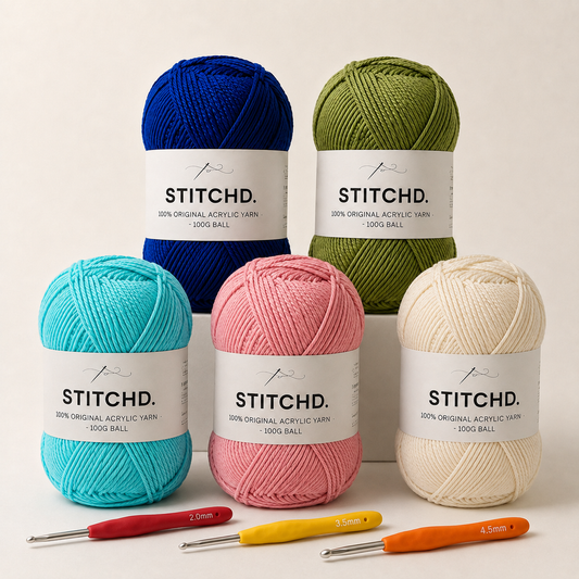Five balls of Stitchd. acrylic yarn in blue, green, turquoise, pink, and cream, arranged on a light beige background with four crochet hooks in front.