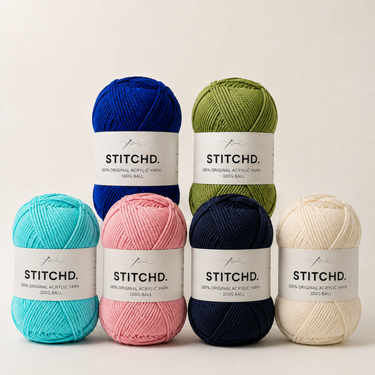 Six balls of Stitchd yarn in assorted colors (blue, green, turquoise, pink, navy, and cream) arranged in two rows against a clean, off-white background.