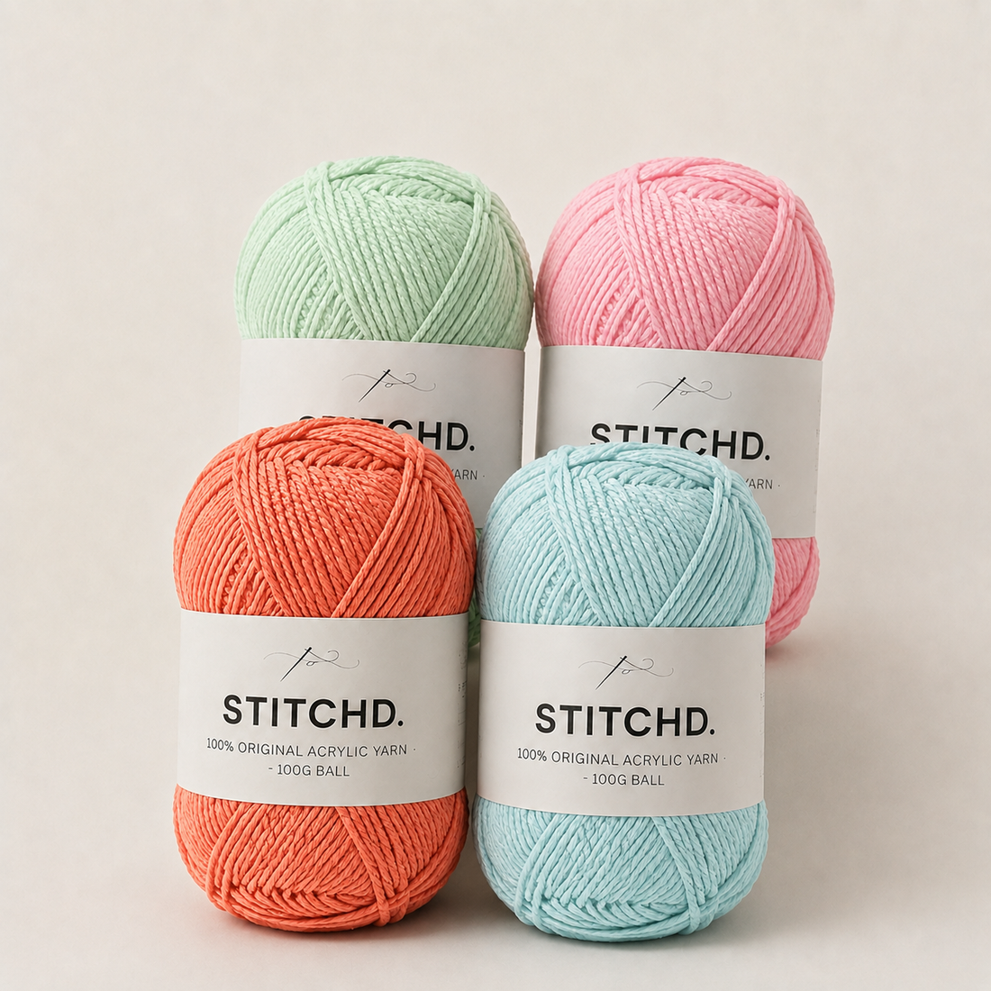 Four balls of Stitchd. acrylic yarn in pastel mint green, coral orange, light blue, and soft pink, arranged in a 2x2 grid on a light beige background.