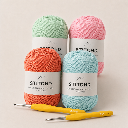 Four balls of Stitchd. acrylic yarn in mint green, coral orange, light pink, and light blue, arranged with two yellow crochet hooks on a warm off-white background.